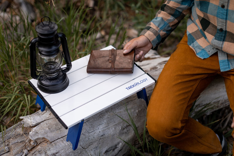 Person sitting on a small wooden table outdoors with a lantern and wallet.